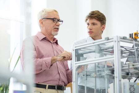 Pleasant interaction. Cheerful senior man communicating with his intern while printing 3D models and...