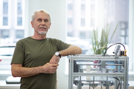 Brand new device. Upbeat senior man posing near a 3D printer and smiling while leaning his elbow on itの写真素材