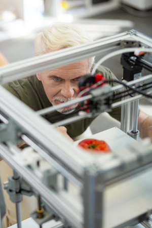 Interesting process. Charming senior engineer leaning closely to the 3D printer and watching a...