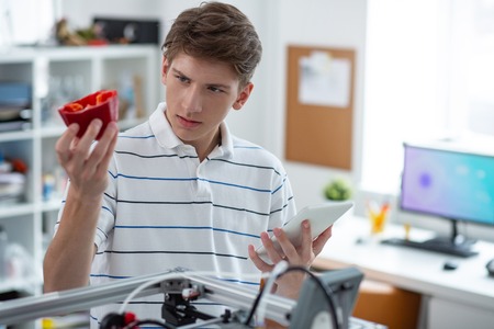 Well done. Pleasant young engineer holding a red bell pepper model and examining it carefully,...