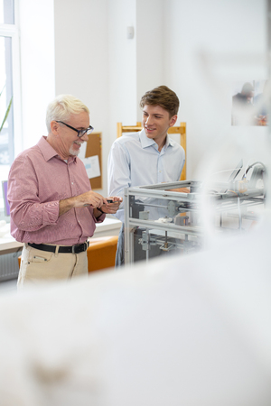 Great mood. Cheerful young man and his senior supervisor joking and smiling while creating models with 3D printerの写真素材