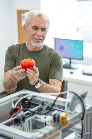 Fantastic result. Cheerful senior man holding a real-like printed red tomato and showing it to his colleagues in the roomの写真素材
