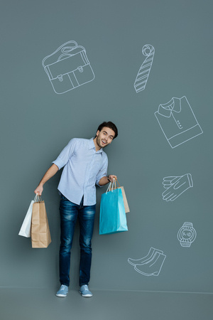 New clothes. Positive emotional young man smiling happily and feeling excited while holding colorful paper bags with his new clothes from the shopping mallの写真素材