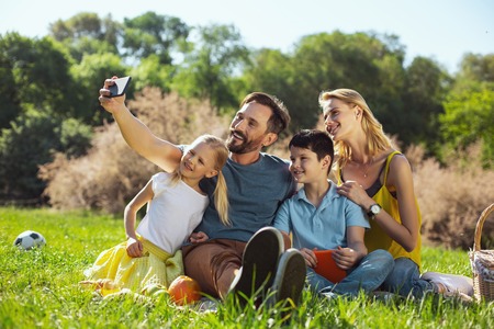 Say cheese. Inspired well-built man smiling and taking selfies with his family in the open airの写真素材