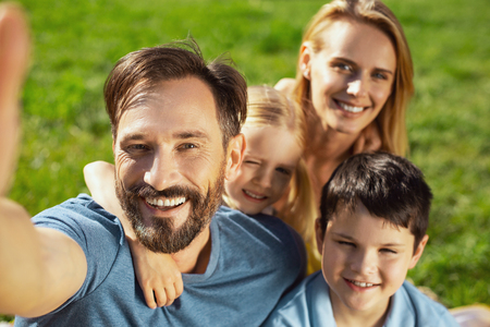 Selfie time. Joyful well-built man smiling and taking selfies with his family in the open airの写真素材