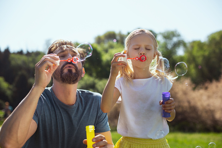 Happy kid. Alert bearded father blowing soap bubbles with his daughter while relaxing in the parkの写真素材