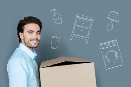 Moving. Positive handsome man holding a carton box and smiling while moving to a new flatの写真素材