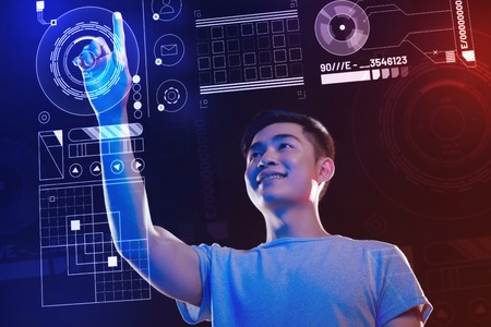 Futuristic technologies. Enthusiastic cheerful software developer smiling and looking interested while touching the round icon on the transparent screen of a futuristic computerの写真素材