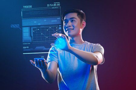 Enthusiastic web developer. Positive emotional web developer feeling happy while standing in his modern office with a tiny gadget in his hand and making a holographic imageの写真素材