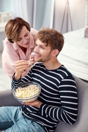 Favourite food. Joyful positive man eating popcorn while enjoying the attention of his motherの写真素材