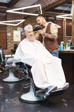 On finish. Pleased pensioner keeping smile on face while sitting on cozy armchair in barbershopの写真素材
