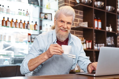 Having pause. Joyful bearded male person holding cup in right hand, looking straight at cameraの写真素材