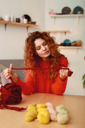 Crochet thread. Appealing ginger-haired girl wearing bright red sweater holding thick maroon crochet threadの写真素材
