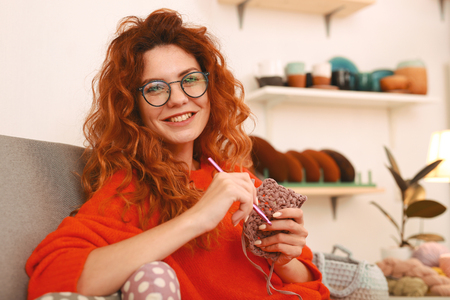 Broad smile. Dark-eyed appealing girl wearing black glasses smiling broadly while knitting clothes for little babies in workshopの写真素材