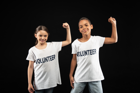 Our team. Cheerful adorable little volunteers wearing white shirts and showing how strong they areの写真素材