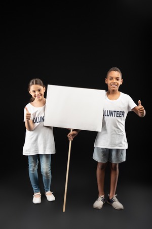 Great job. Pleased adorable little volunteers wearing white shirts and holding a white tableの写真素材