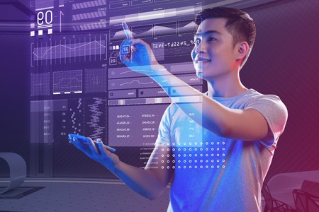 Entering new data. Clever positive web developer having a smartphone in his hand and smiling while touching the hologram and analyzing new dataの写真素材