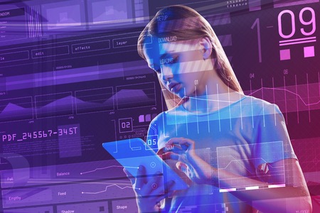 Careful student. Serious young student standing in front of a transparent screen and looking attentively at the screen of a modern tablet in her handsの写真素材