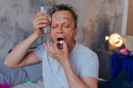 Alcohol intoxication. Tired mature man holding water and swallowing pillの写真素材