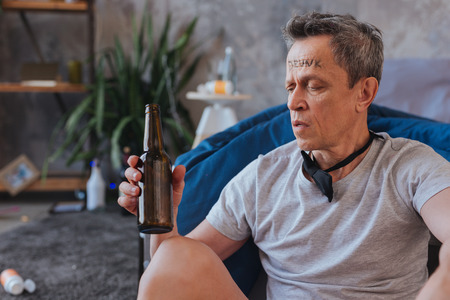 Fluid loss. Earnest mature man sitting on floor and staring at bottleの写真素材