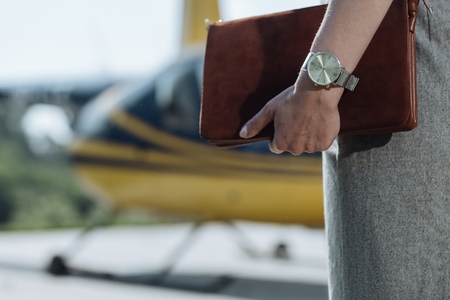 Stylish accessories. The close up of a delicate female hand with a watch on it holding a fashionable bag during the visit to a helipadの写真素材