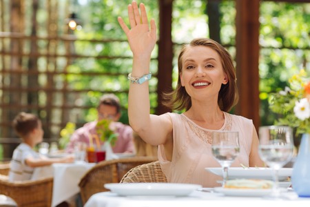 Lifting hand. Smiling dark-eyed woman wearing stylish beige dress lifting her hand calling waitressの写真素材