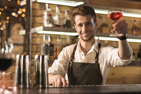 There you go. Pleasant young barman standing behind the bar counter and handing a cocktail to the customer while smilingの写真素材