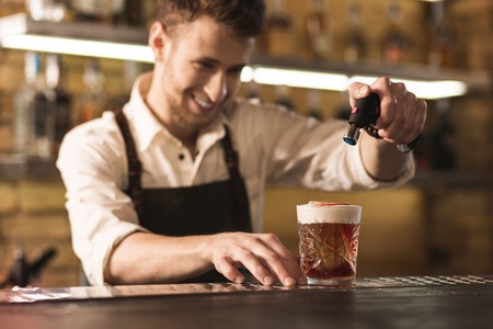 Special equipment. Joyful professional barman standing behind the bar counter and using a culinary torch to burn the sugar on his cocktailの写真素材