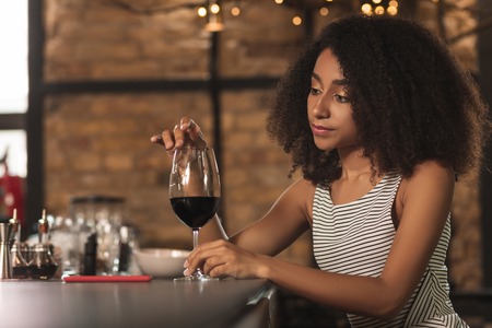 Relaxing in bar. Beautiful young woman touching the rim of a glass of a red wine with her finger while sitting in the barの写真素材