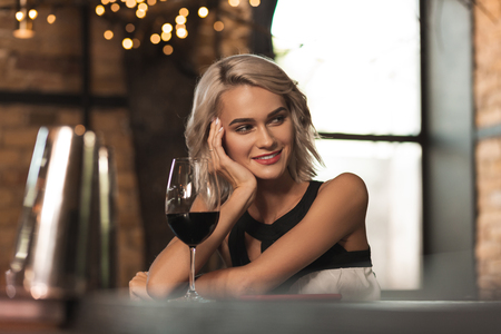 Welcoming smile. Charming blonde woman sitting at the bar counter, drinking wine and smiling at someone while resting her cheek on her palmの写真素材