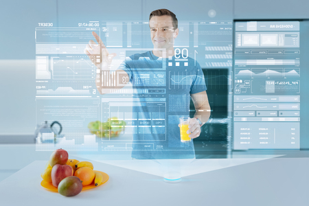 Easy usage. Cheerful handsome man holding a glass of fresh juice while and smiling while standing in a modern kitchen and touching a transparent projection over the countertopの写真素材