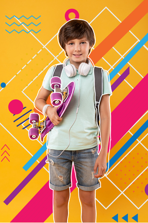 Active rest. Positive relaxed teenager wearing headphones and looking glad while standing with a bright skateboard in his handsの写真素材