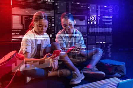 Comparing smartphones. Curious modern children having a good time after school while sitting on the floor and looking at their modern gadgetsの写真素材