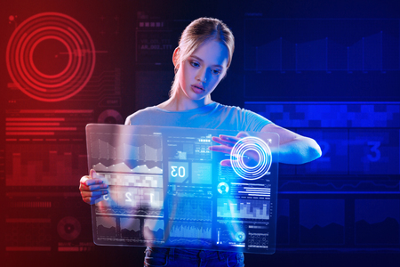 Transparent gadget. Calm clever teenage girl carefully holding a transparent device and touching the icons on the screen of it while preparing her school projectの写真素材