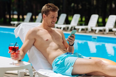 Reading message. Handsome muscle man reading message on his blue smart phone while getting suntan