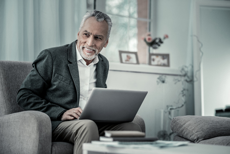 Have idea. Handsome male keeping smile on his face, holding laptop on knees and looking asideの写真素材