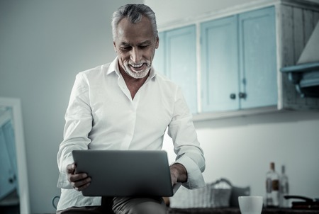 Work at home. Pleased mature male bowing head while looking at screen of his laptopの写真素材