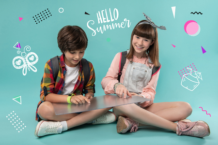 Hello summer. Positive friendly teenagers sitting together with a big transparent device and looking at the screen of it while waiting for their journeyの写真素材
