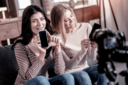 Cutting-edge phones. Happy pretty bloggers holding their phones while recording a new video for their blogの写真素材
