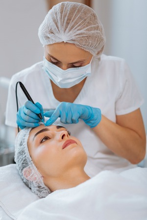 Professional therapy. Serious skilled dermatologist holding a special device while removing a mole