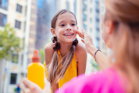 Spf protection. Delighted happy girl standing in front of her mother while having sunscreen on her...