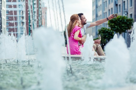 Our memory. Joyful delighted couple sitting near the fountain while taking selfies togetherの写真素材