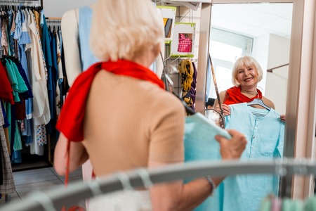 Incredible look. Pleased fashionable elderly woman looking at her reflection in shopping store mirror while admiring her new stylish dressの写真素材