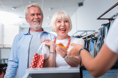 Mission done. Delighted stylish elderly couple standing in front of cashier desk while passing a...