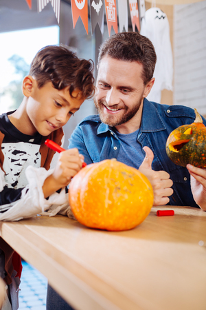 Complimenting pumpkin. Loving father complimenting bright Halloween pumpkin of his cute son wearing skeleton costumeの写真素材