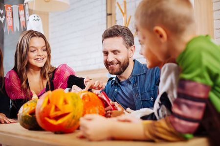 Joining children. Handsome bearded father feeling happy while joining his children coloring and decorating pumpkinsの写真素材