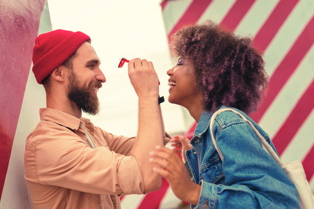Holding glasses. Positive bearded man smiling cheerfully while standing in front of his happy girlfriend and holding her red glassesの写真素材
