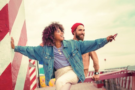 Enjoying view. Emotional positive young woman sitting on the hand rail and pointing to the distance while showing the view to her smiling boyfriendの写真素材