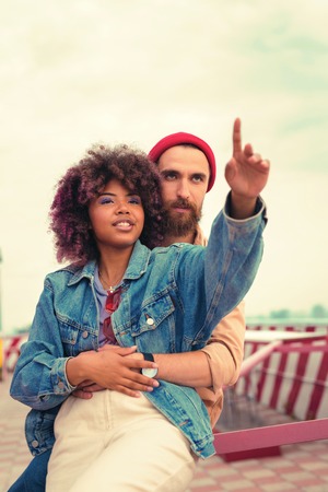 Pointing at the sky. Calm handsome young man hugging his smiling girlfriend and looking at the skyの写真素材