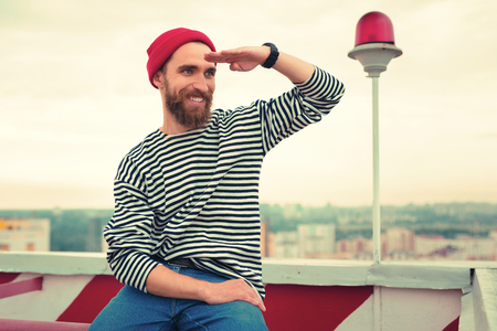 Looking glad. Cheerful bearded young man wearing red hat and putting his hand to the face while looking into the distance and smilingの写真素材